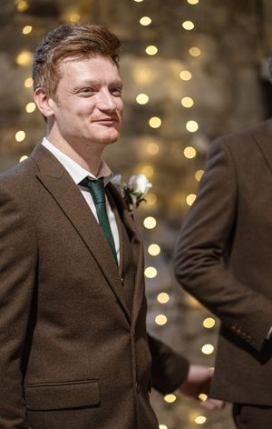 A person wearing a brown suit and green tie smiles at a formal event with twinkling lights in the background.
