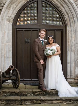 A newlywed couple poses in front of a grand arched wooden door at a historic stone church with an antique cannon nearby.