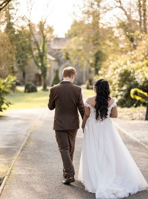 A couple in wedding attire walks along a scenic outdoor path during golden hour.