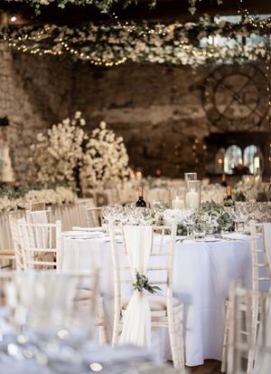 Elegant wedding reception setup with white chairs and round tables decorated with floral centerpieces under twinkle lights.