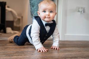 Young toddler in formal attire crawls across hardwood floor at wedding celebration.