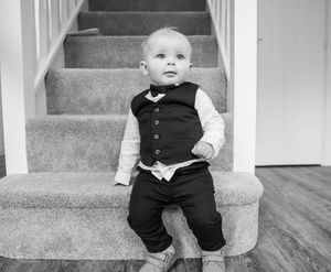 Small child wearing formal black vest and white shirt sitting on carpeted stairs in black and white photo.
