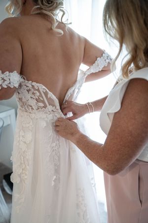 Someone helping to fasten the back of an ornate lace wedding dress with delicate floral details.