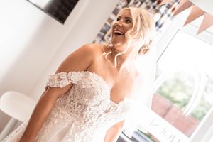 A bride wearing an elegant lace wedding dress with ruffle details stands in a bright room during her wedding preparations.