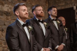 Groomsmen in dark suits stand in line against a rustic stone wall backdrop.