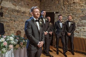 Groomsmen in dark suits lined up against rustic stone wall during wedding celebration.