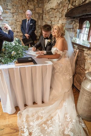 A couple signs their marriage certificate at a rustic venue with stone walls and elegant floral decorations.