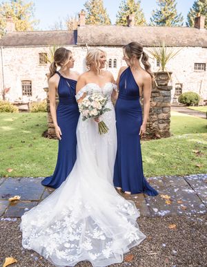 Bridesmaids in navy blue dresses pose with bride's lace gown train outdoors at stone venue.