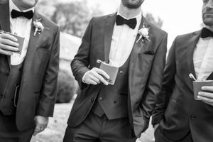 Wedding party members in formal attire sharing drinks and laughter outdoors.
