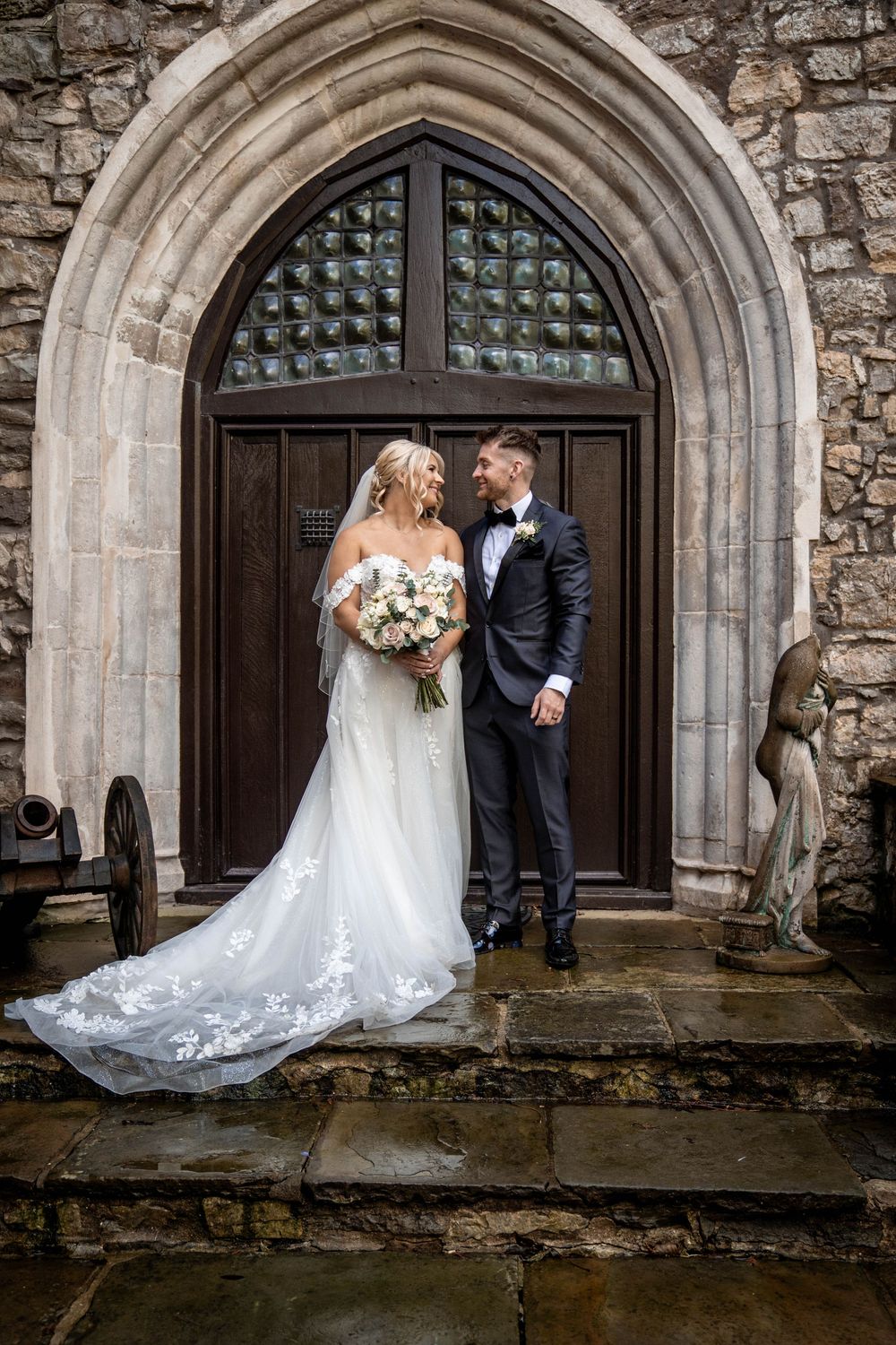 A romantic wedding couple stands in front of a grand stone church archway with ornate Gothic architectural details.