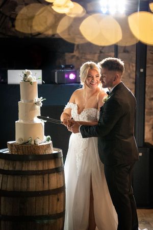 A wedding couple share an intimate moment next to their elegant white wedding cake displayed on a rustic wooden barrel.