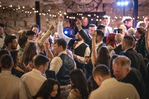Wedding guests dance and mingle at a rustic stone-walled venue illuminated by string lights overhead during an evening reception.