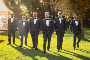Wedding party in black tuxedos walking together on a sunny lawn with golden hour lighting in the background.