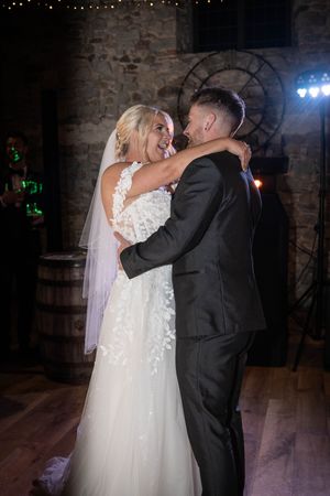Bride in flowing white gown and groom in dark suit share a romantic first dance at rustic brick venue.