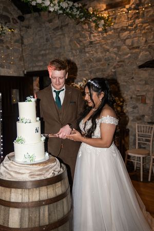 A couple cuts their elegant white wedding cake displayed on a rustic wooden barrel at a stone-walled venue.