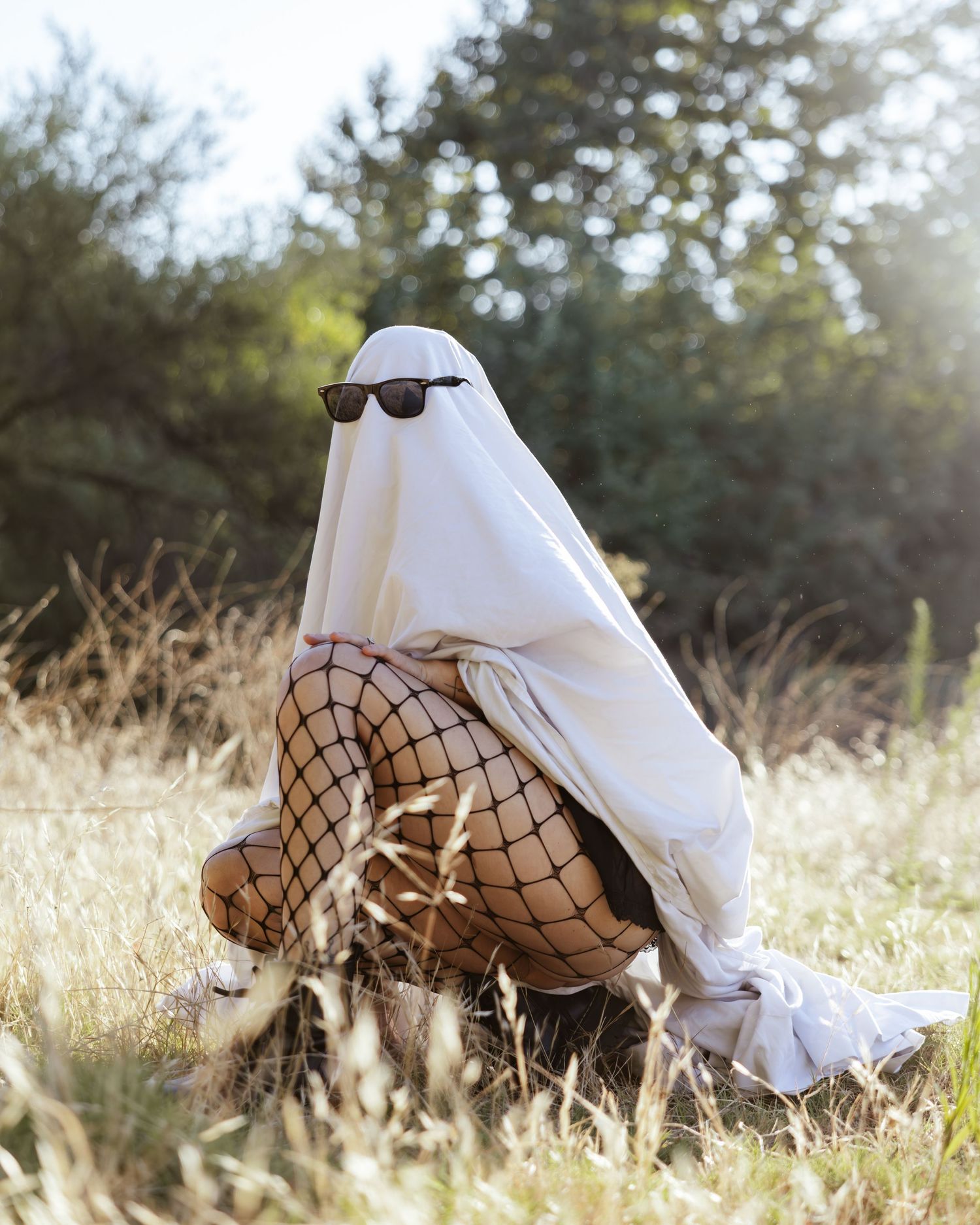 Person in white fabric and fishnet stockings posing dramatically on sunlit grass with artistic flair.
