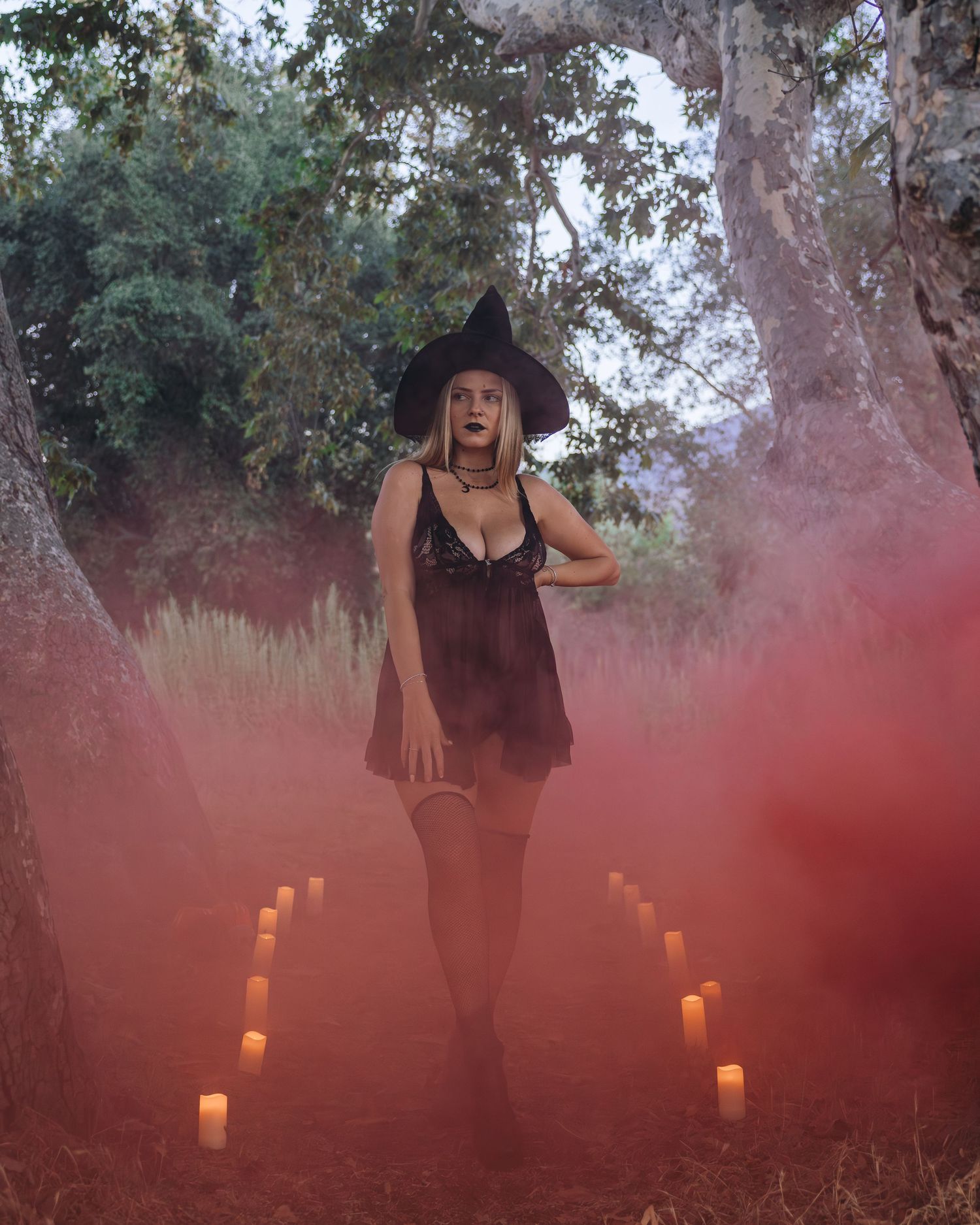 A mystical scene with glowing candles and red smoke around a figure in a black dress and hat in a forest setting.