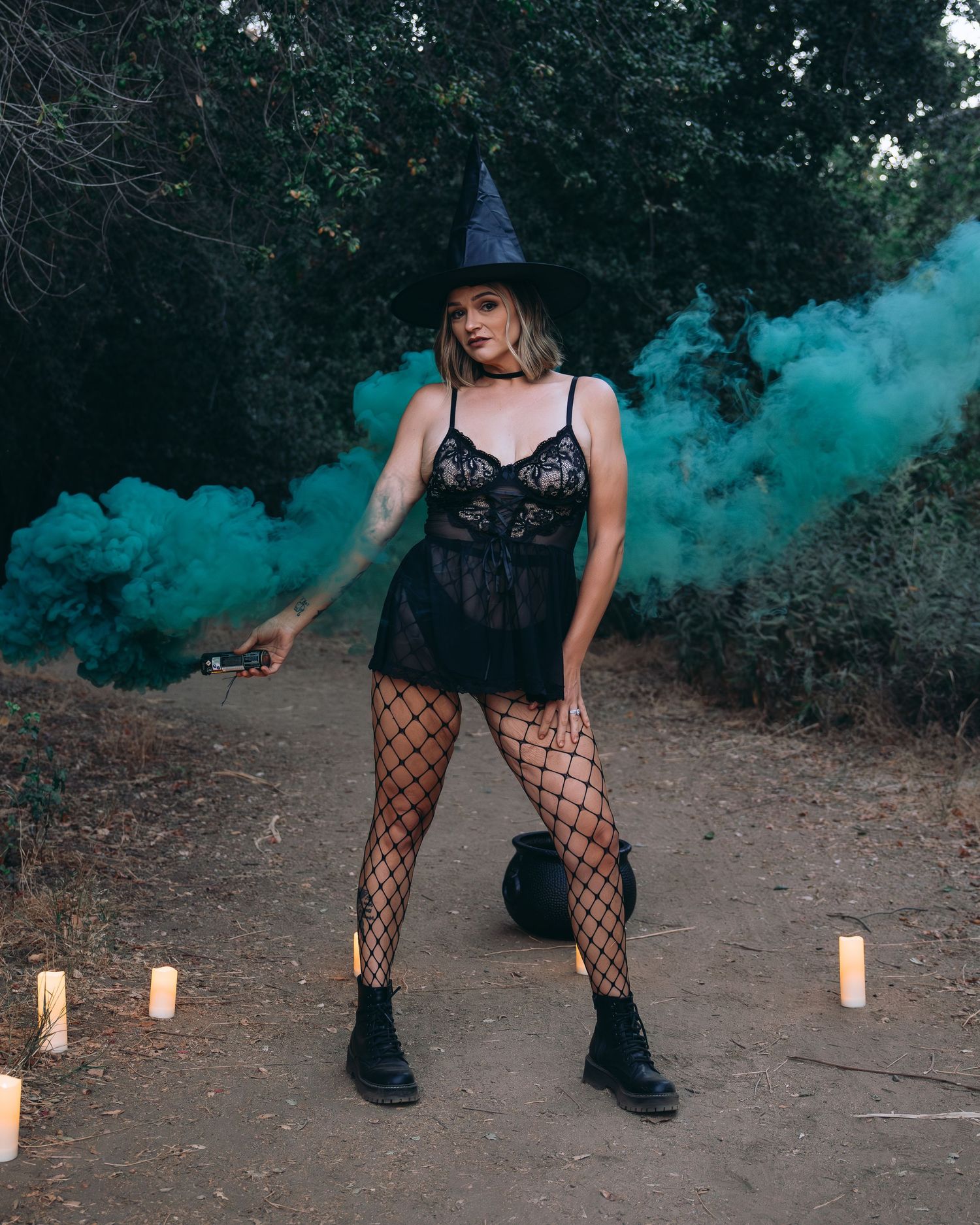 Gothic fashion photoshoot in a dark forest setting with turquoise smoke and black boots on a dirt path.
