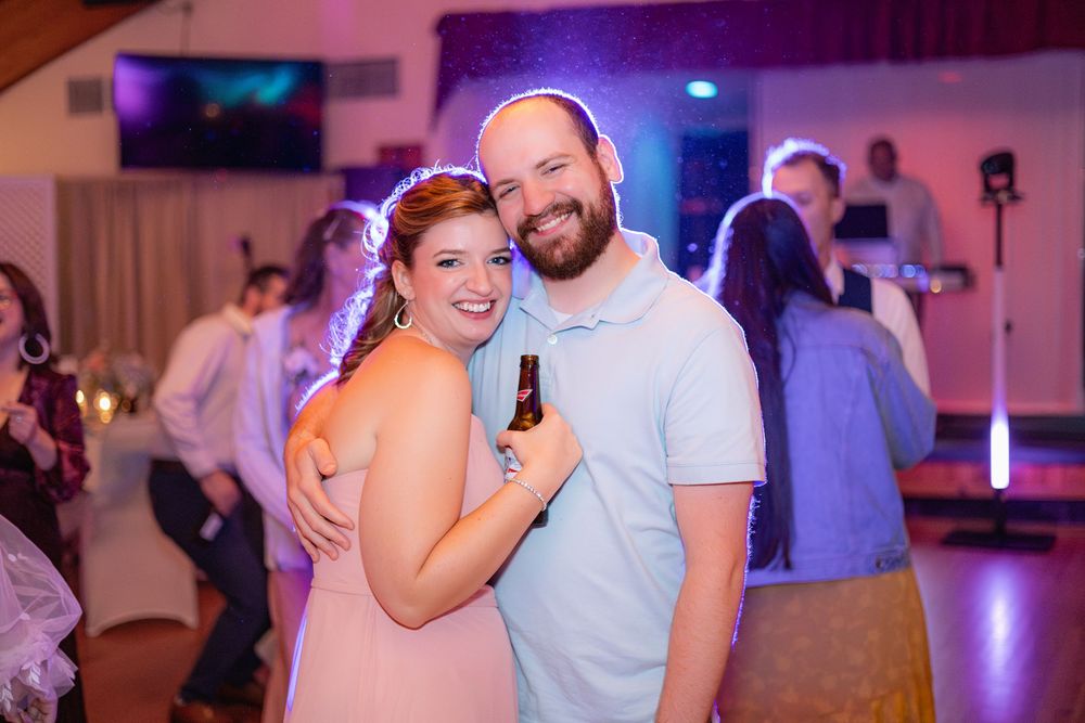 A couple dances at a wedding reception with colorful pink and purple ambient lighting in the background.