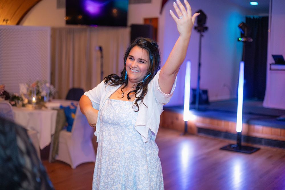 A person in a white lace dress raises their arm while dancing at a reception venue with purple lighting.
