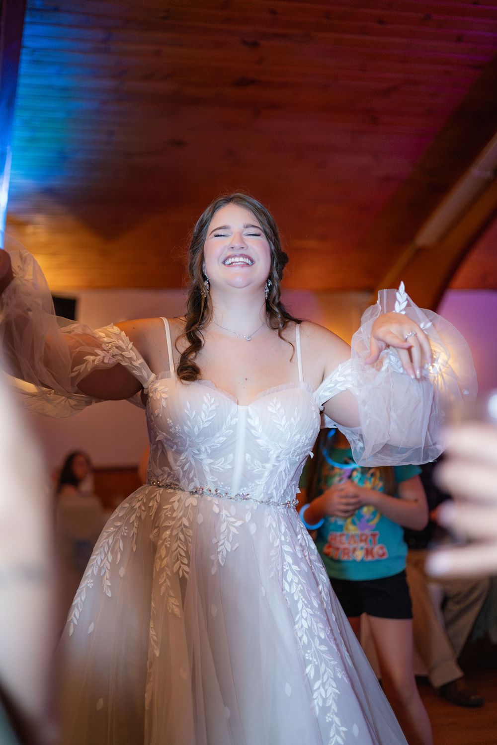 A person in a white lace wedding dress dances joyfully at a reception with colorful lighting.