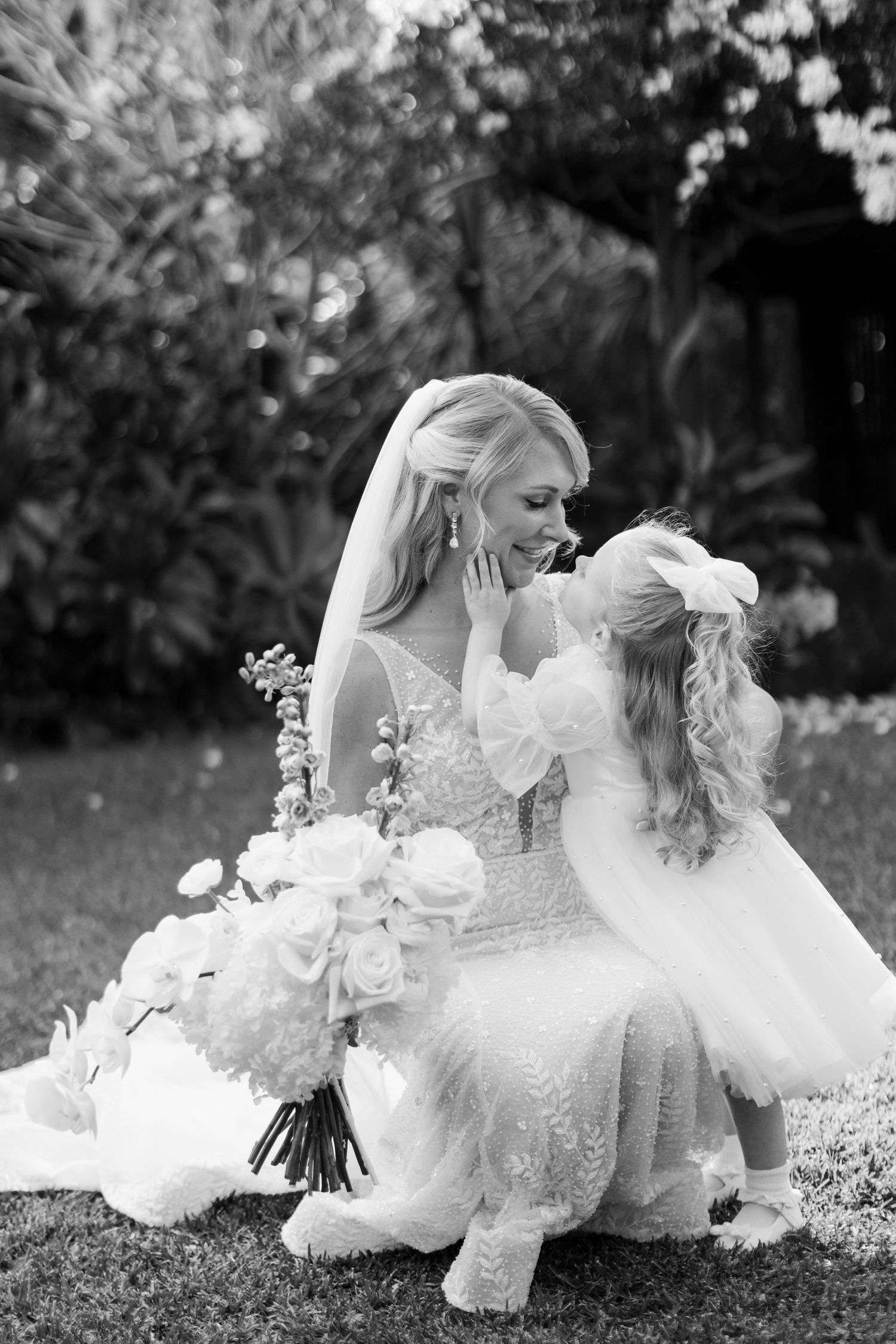 A tender black and white photograph of a bride in a lace gown sharing a sweet moment with her flower girl in a garden.