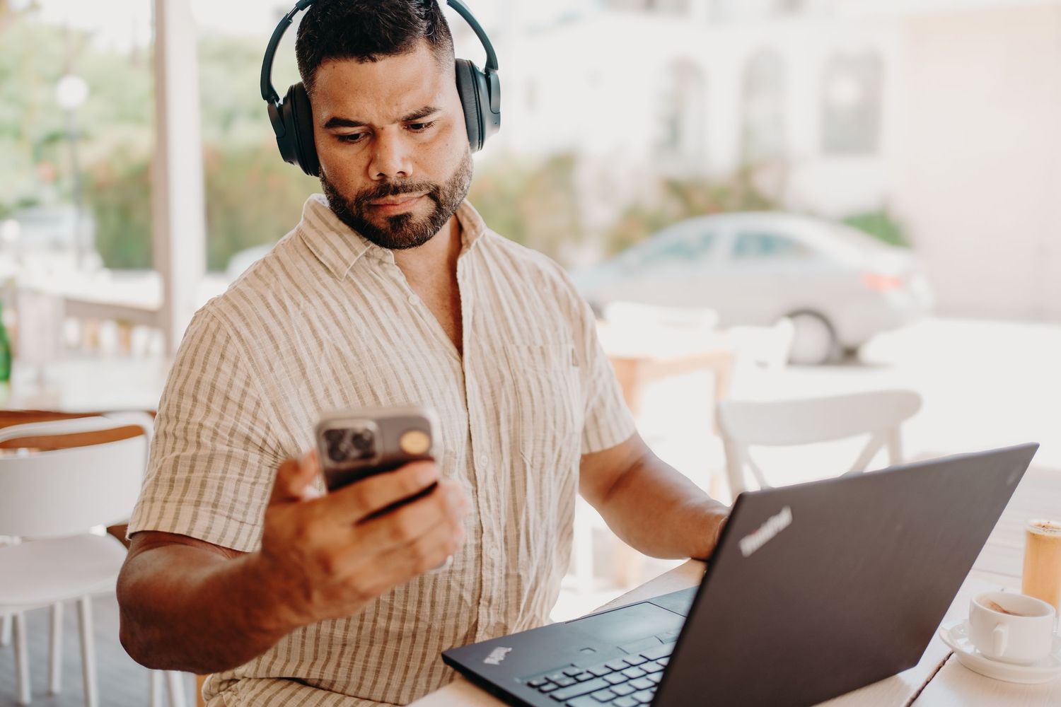 Remote worker in beige striped shirt using laptop and headphones while checking smartphone at home office setup. Photoshoot Puerto Morelos.