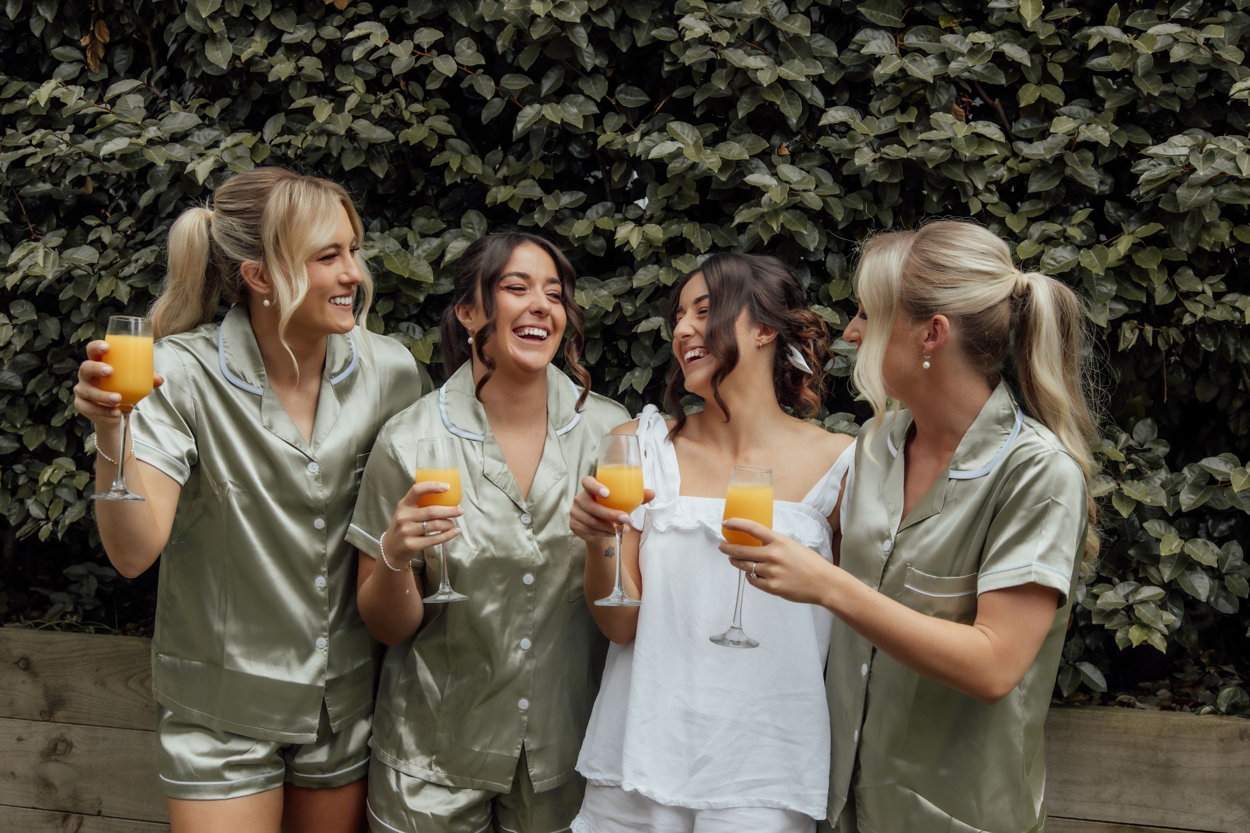 Friends laughing and toasting with drinks at an outdoor gathering wearing coordinated olive and white outfits.