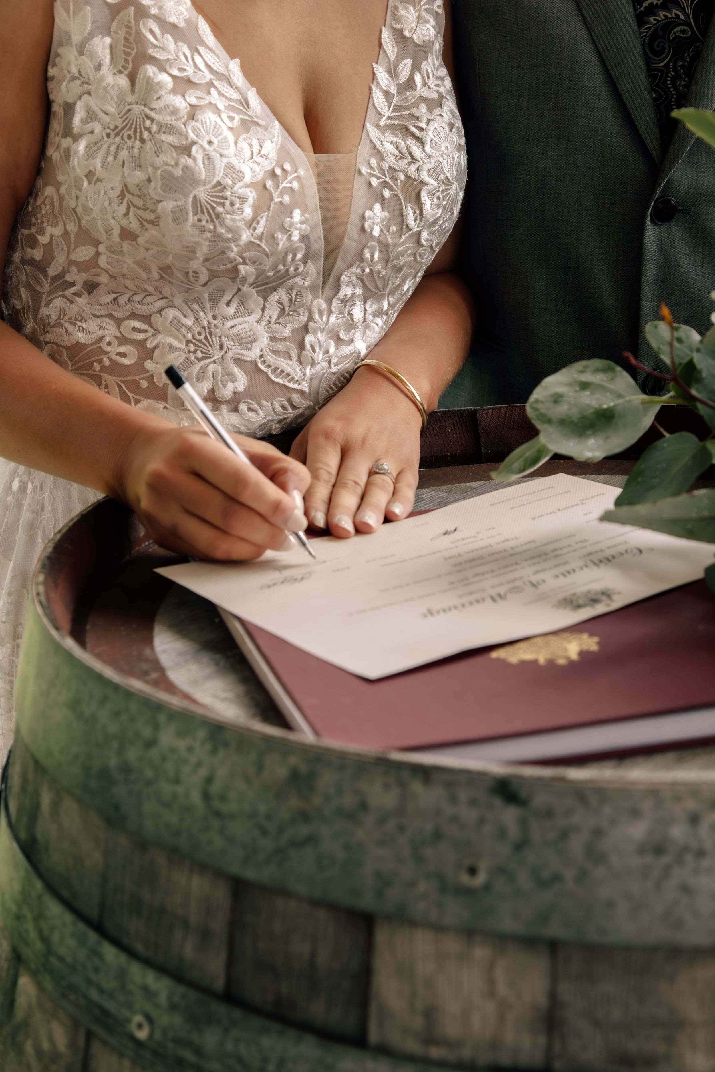 Close-up shot of someone signing documents on a rustic wine barrel with lace wedding dress details visible in frame.