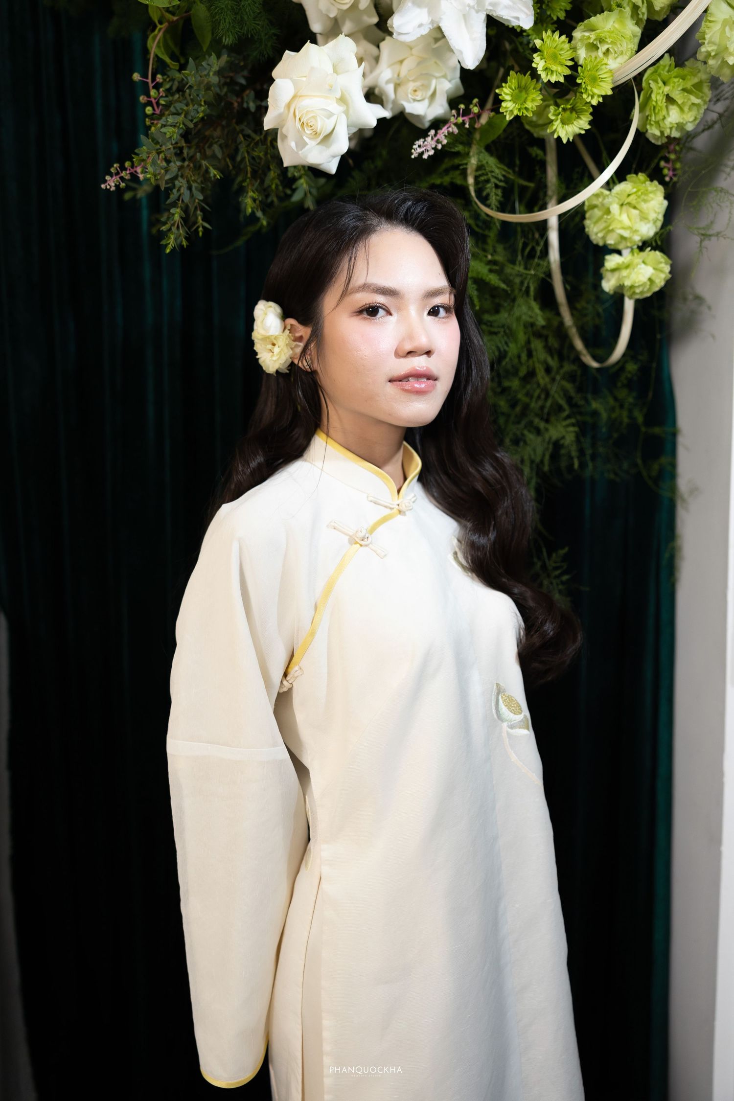 Young Vietnamese woman in elegant cream ao dai standing before a lush floral arrangement and green drapes.