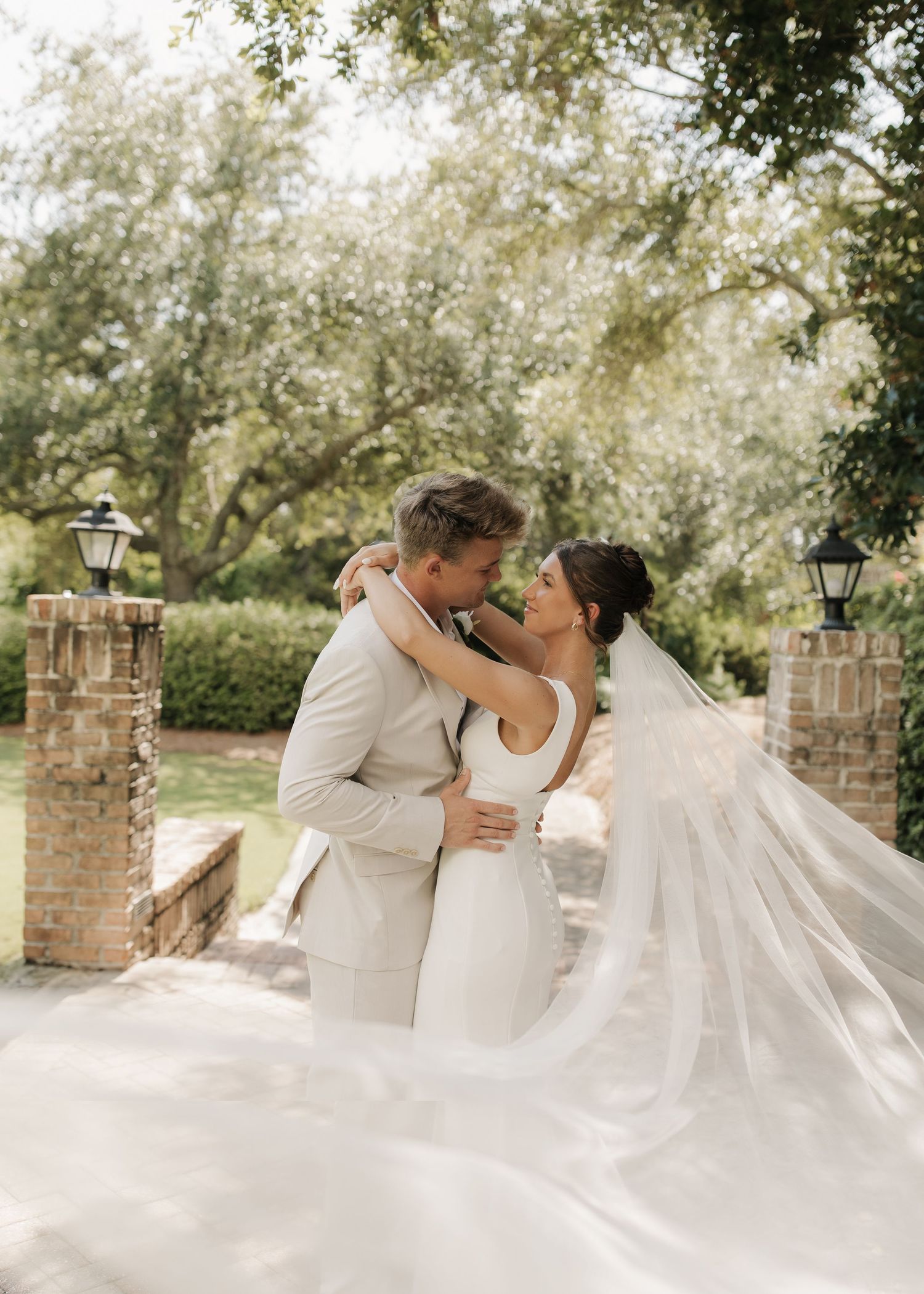 A romantic wedding couple shares a series of kisses under a lush tree canopy on a garden path.