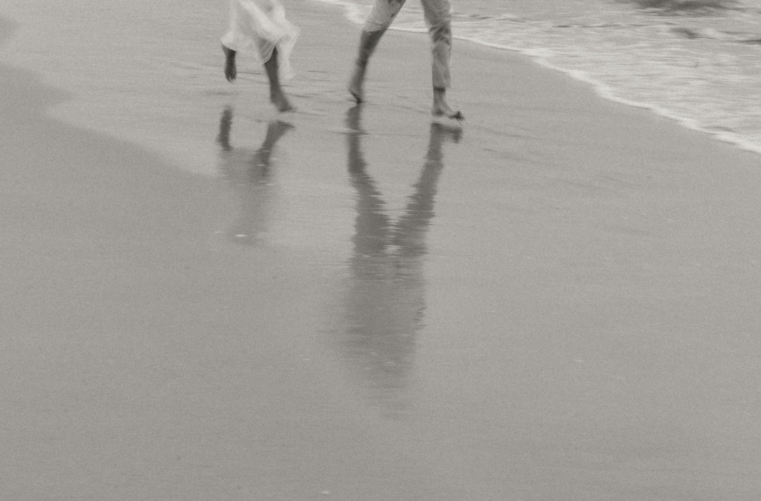 Two people stand on a wet reflective beach at sunset casting long shadows in the sand while waves roll in.