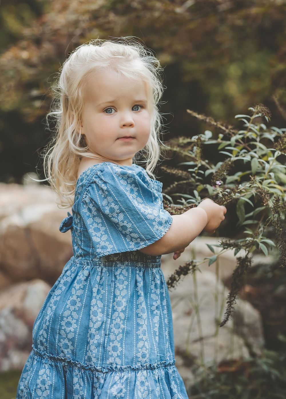 A small child in a blue floral dress stands in a garden reaching toward plants and flowers.