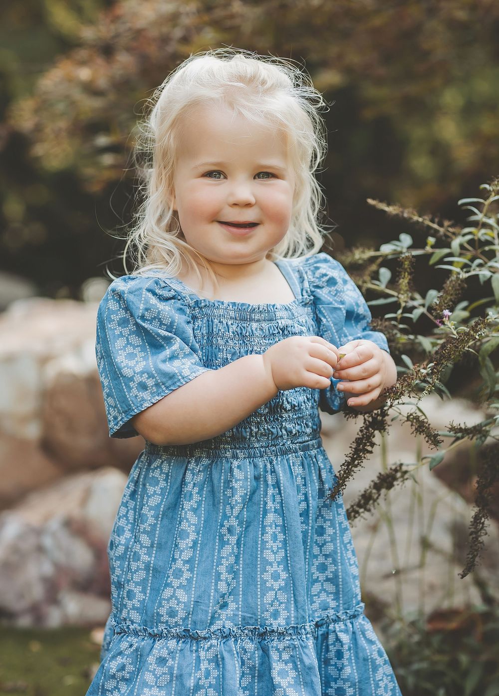 A young child in a blue floral dress reaches toward tree branches in a dreamy outdoor garden setting.