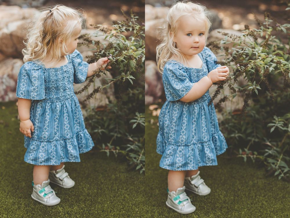 Small child wearing a blue floral dress and white shoes stands in a garden setting with greenery in the background.