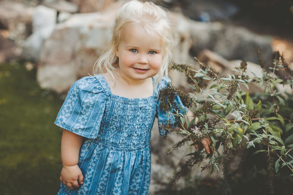 A child wearing a blue floral dress stands in a garden setting during golden hour lighting.