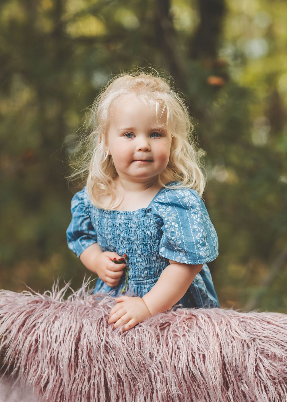 A toddler in a blue smocked dress leans over a pink furry blanket outdoors during a photo session.
