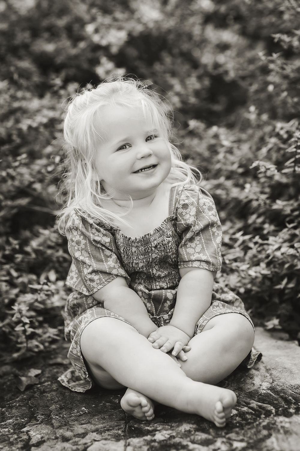 Black and white portrait of a young child sitting outdoors in a floral dress with light hair and a joyful smile.