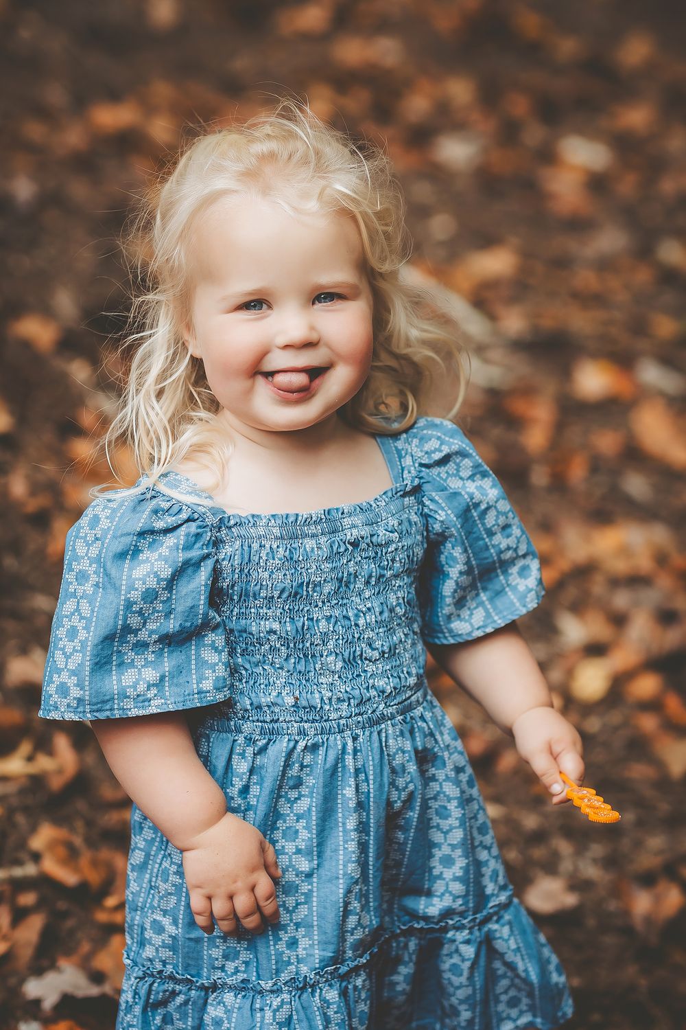 A small child wearing a blue ruffled dress stands among autumn leaves while smiling and holding something yellow.