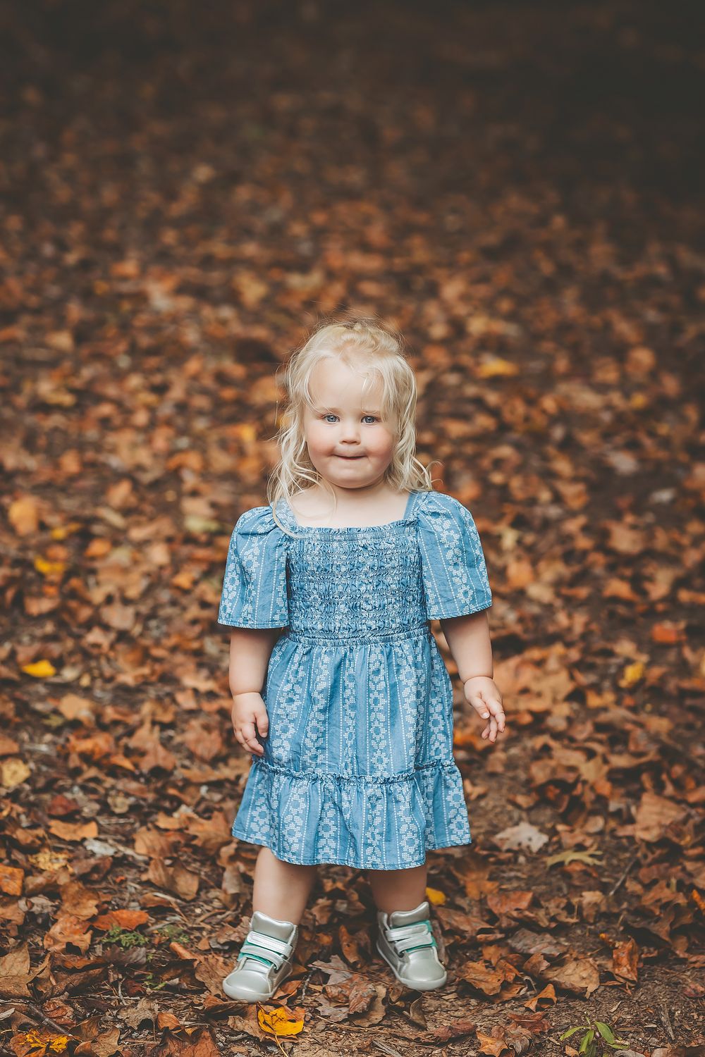 A small child in a blue dress sits and plays among scattered autumn leaves on the ground.