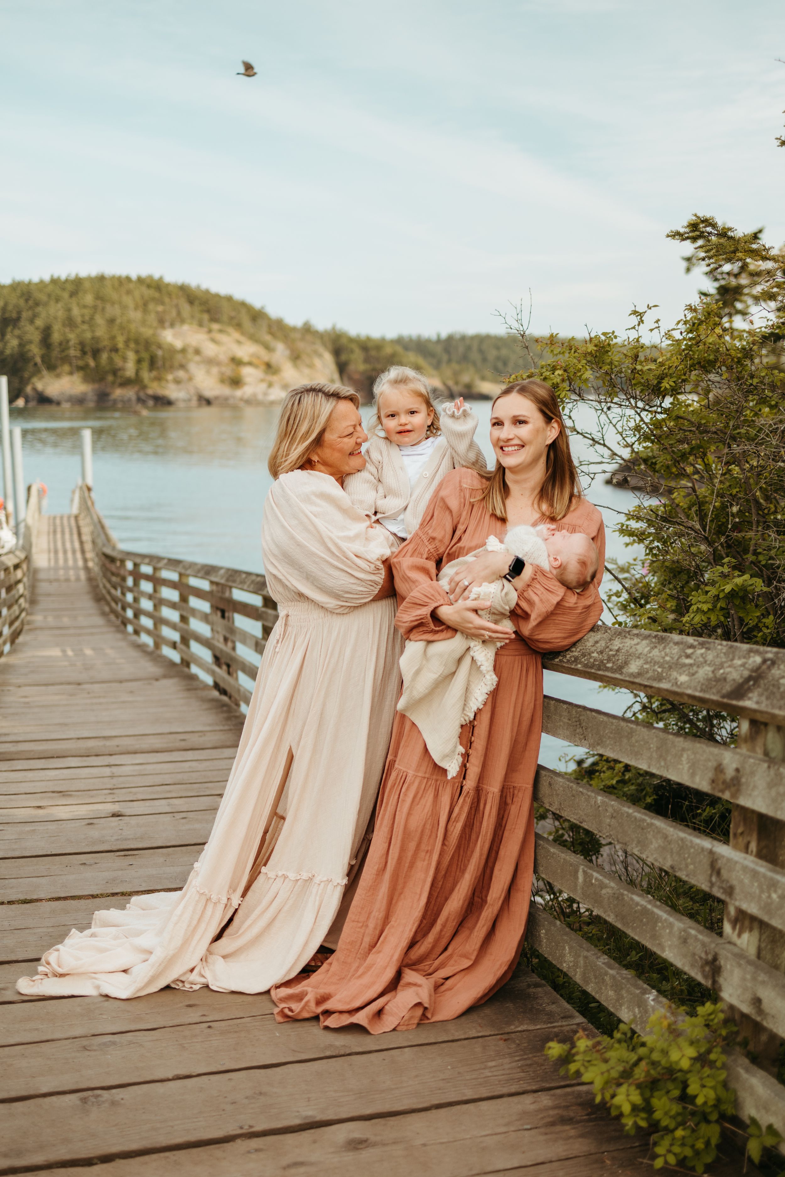 A group wearing flowing dresses in muted tones sits together on a wooden boardwalk overlooking a scenic coastal landscape.