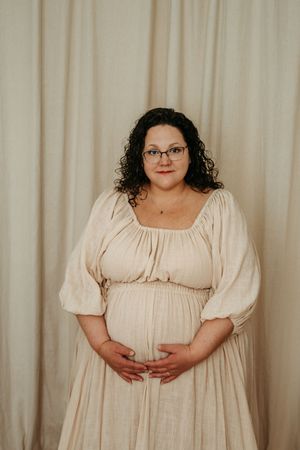 A pregnant person in a cream-colored empire waist dress with puff sleeves poses against a light backdrop.
