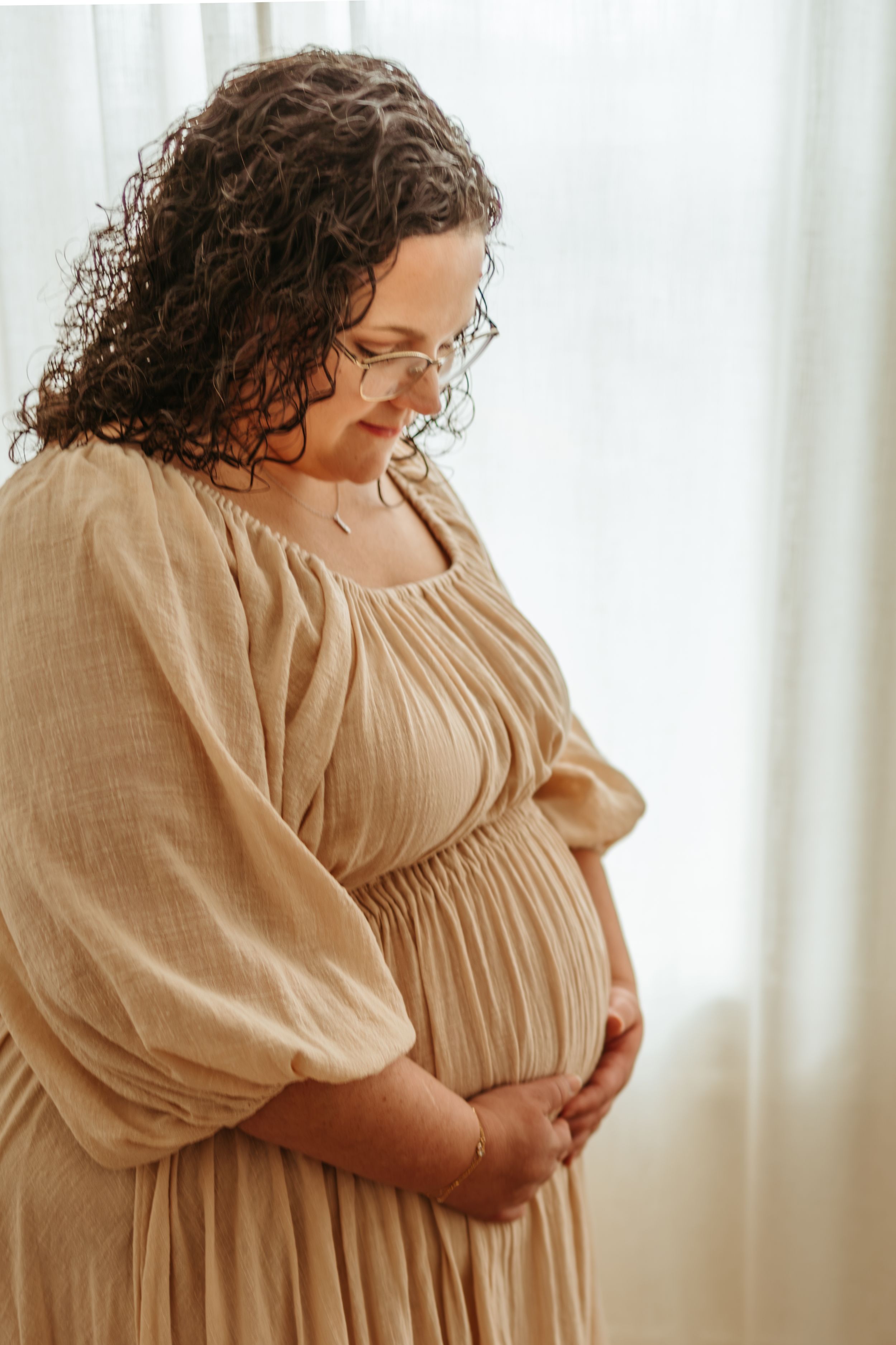 A person in a beige linen dress stands by a window in a series of intimate maternity photos.