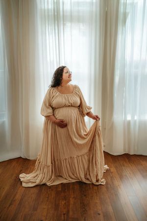 Four artistic poses of a figure in a flowing beige dress on wooden flooring with sheer white curtains in background.