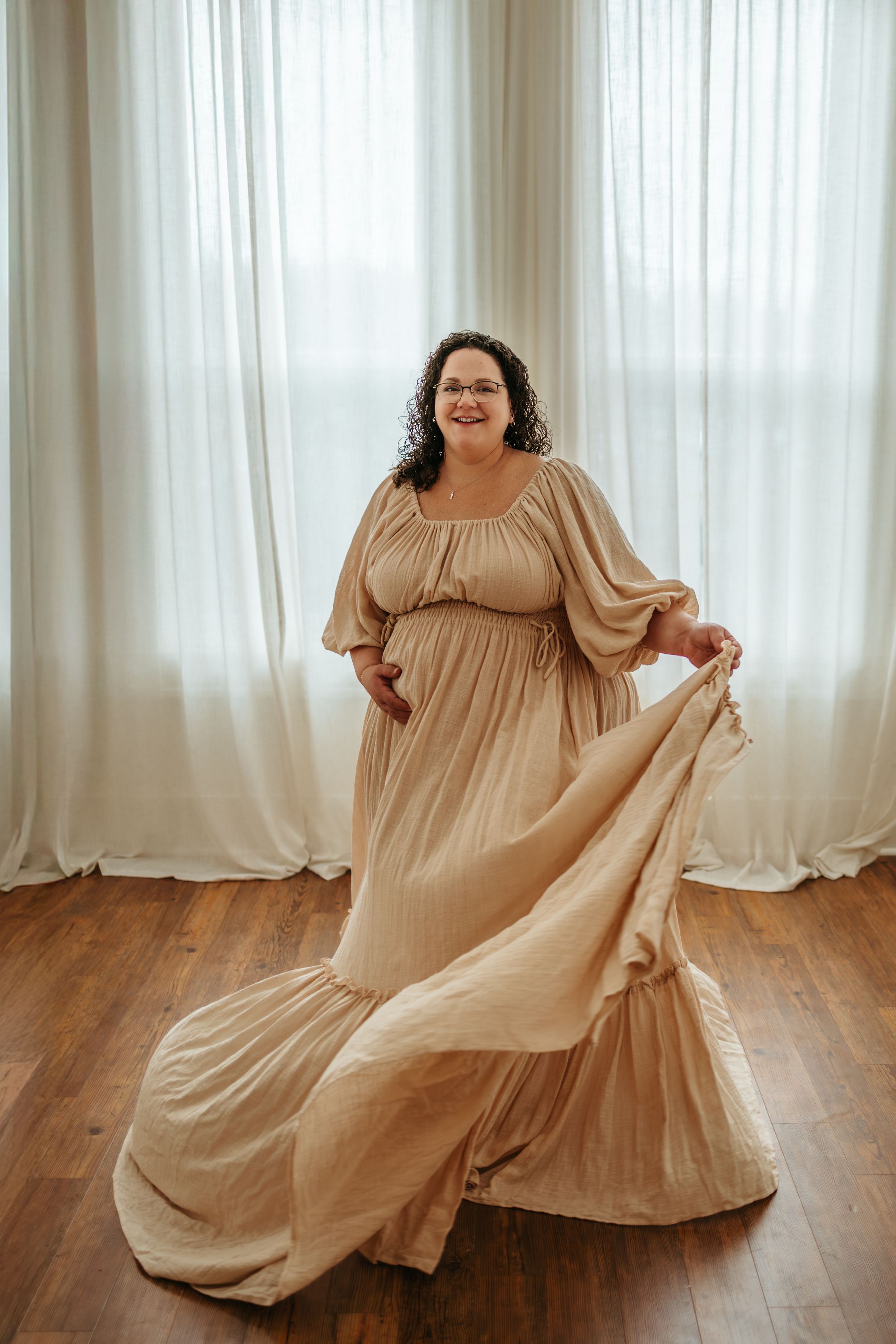 A person in a flowing beige dress twirls gracefully across a hardwood floor in front of sheer white curtains.