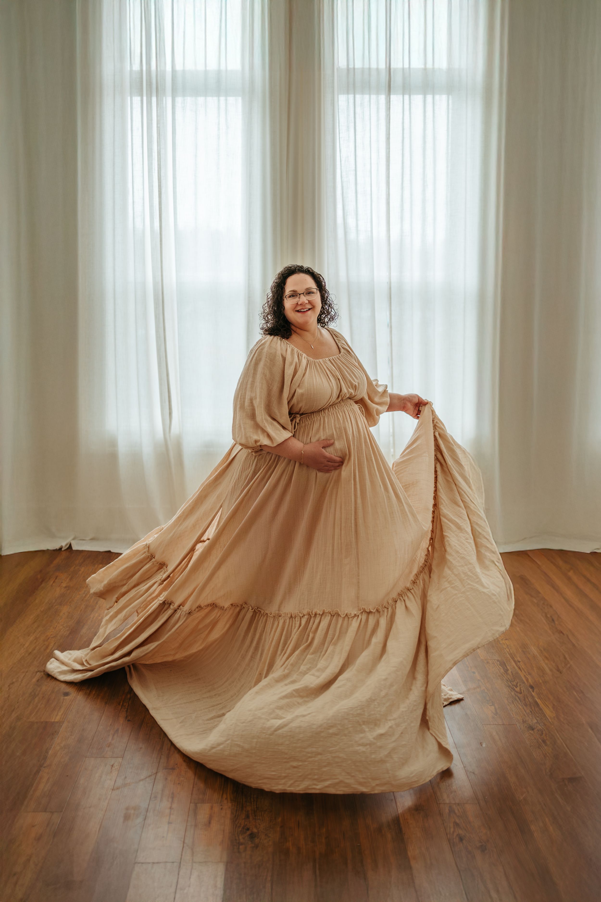 A person in a flowing beige dress twirls gracefully on hardwood flooring in front of sheer curtained windows.