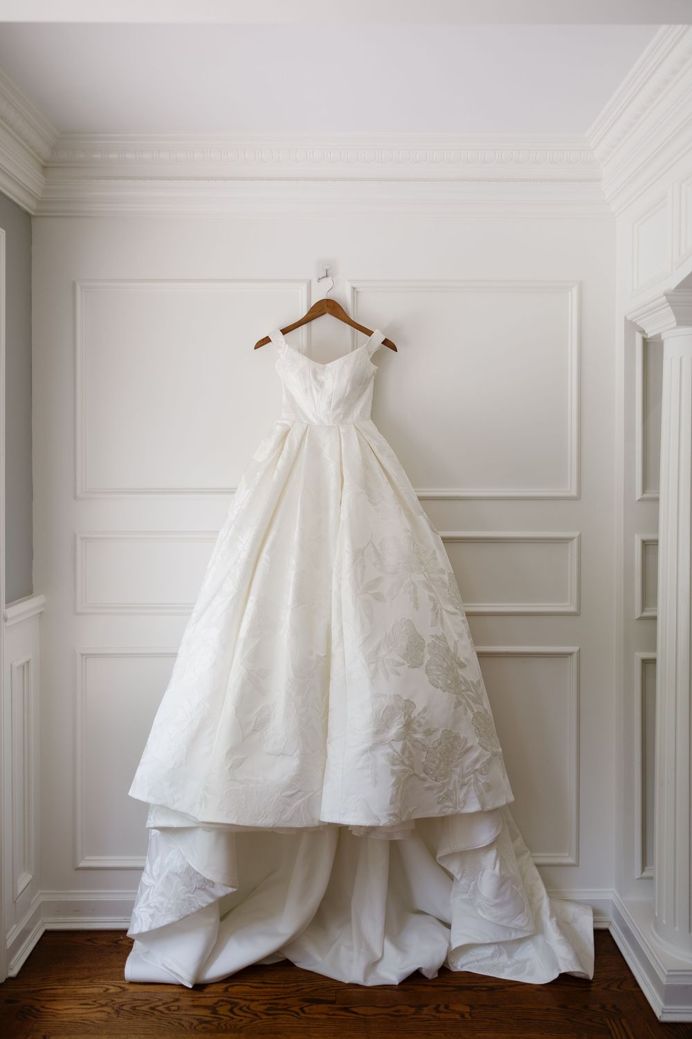 White wedding dresses displayed on hangers against a classic white paneled wall with hardwood floors.
