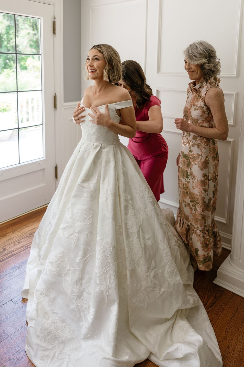 Wedding preparations show a flowing white gown being adjusted in a bright room with hardwood floors.