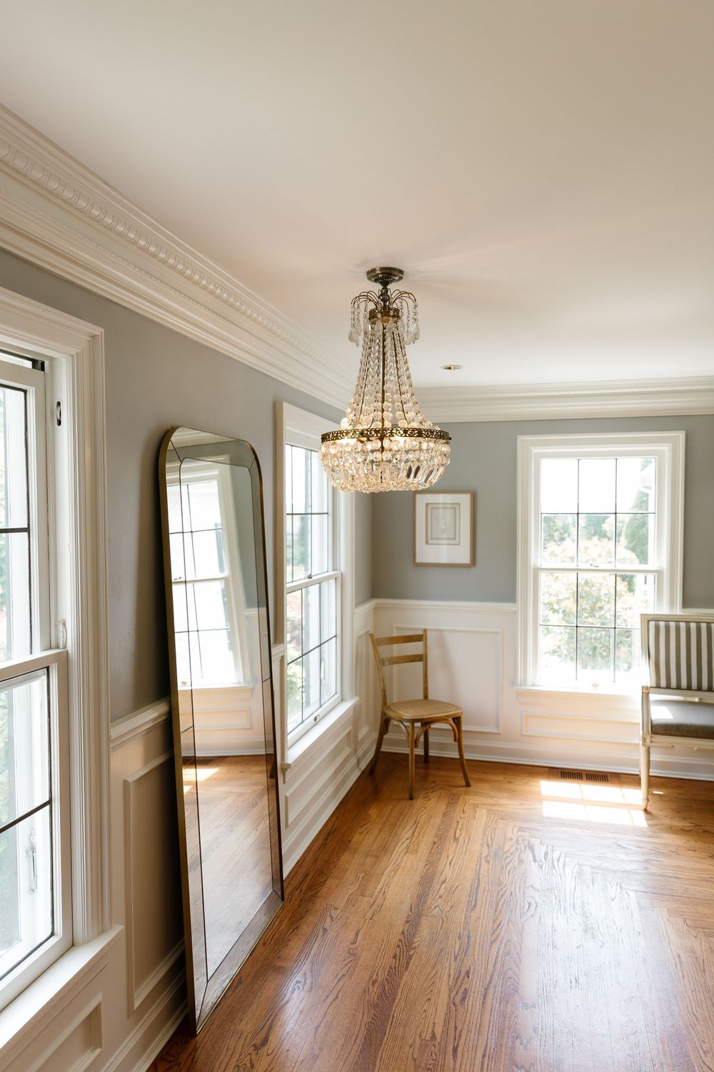 Spacious empty room with hardwood floors, white crown molding, and crystal chandeliers featuring large windows.