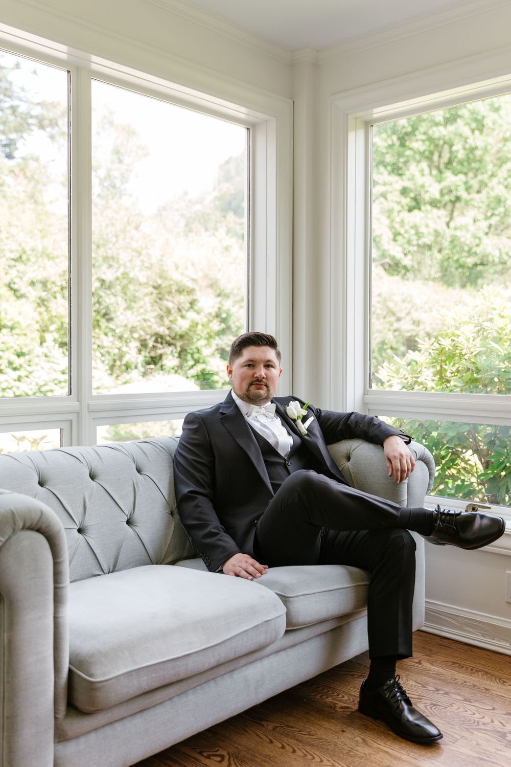 A light gray couch sits in a bright sunlit room with large windows and hardwood floors.